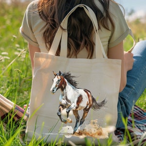 May include: A cream-coloured canvas tote bag with a watercolour painting of a running horse. The horse has a white and brown coat and a flowing black mane. The bag has two handles and is held by a person sitting in a grassy field.