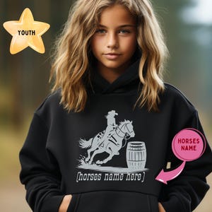 May include: Black hooded sweatshirt with a barrel racing design. The graphic shows a rider on a horse racing around a barrel. The word "YOUTH" is on a gold star. The text "horses name here" is below the graphic.