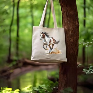 May include: A cream-coloured tote bag displaying a watercolour painting of a running horse. The horse is brown and white with a flowing mane and tail. The background is a blurred image of a forest, with the bag hanging from a tree.