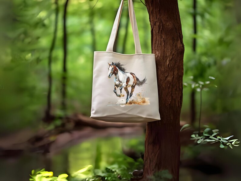 May include: A cream-coloured tote bag displaying a watercolour painting of a running horse. The horse is brown and white with a flowing mane and tail. The background is a blurred image of a forest, with the bag hanging from a tree.