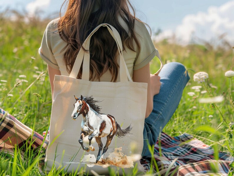 May include: A cream-coloured canvas tote bag with a watercolour painting of a running horse. The horse has a white and brown coat and a flowing black mane. The bag has two handles and is held by a person sitting in a grassy field.
