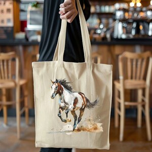 May include: A beige canvas tote bag with a watercolour painting of a running horse. The horse is brown and white with a flowing mane and tail. The bag has long handles and is held by a person.
