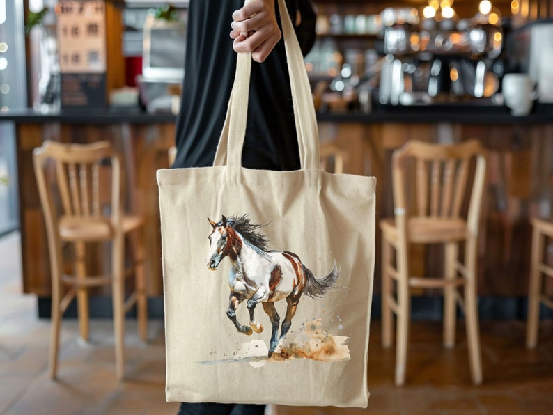 May include: A beige canvas tote bag with a watercolour painting of a running horse. The horse is brown and white with a flowing mane and tail. The bag has long handles and is held by a person.