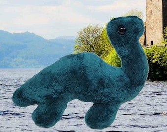 Könnte beinhalten: Ein tealfarbenes Plüschtier in Form des Ungeheuers von Loch Ness, mit langem Hals und kleinem Kopf. Das Spielzeug hat eine weiche, flauschige Textur und ein dunkles Auge. Der Hintergrund zeigt einen See, Berge und ein Schloss.