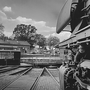May include: A black and white photo of a vintage steam locomotive parked on train tracks. The locomotive is partially visible, with its wheels and engine details in focus. The photo also shows a train station building and a small wooden shed in the background.
