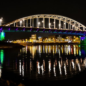Op de afbeelding: Een brug verlicht met paarse en groene lichten 's nachts, die weerspiegeld worden in het water eronder. De brug heeft een stalen boogconstructie en wordt verlicht door straatverlichting.