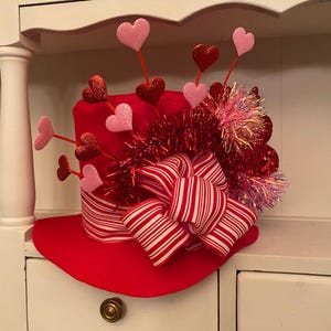 May include: A red top hat with a white and red striped bow, pink and red hearts, and red and pink tinsel. The hat is sitting on a white shelf.