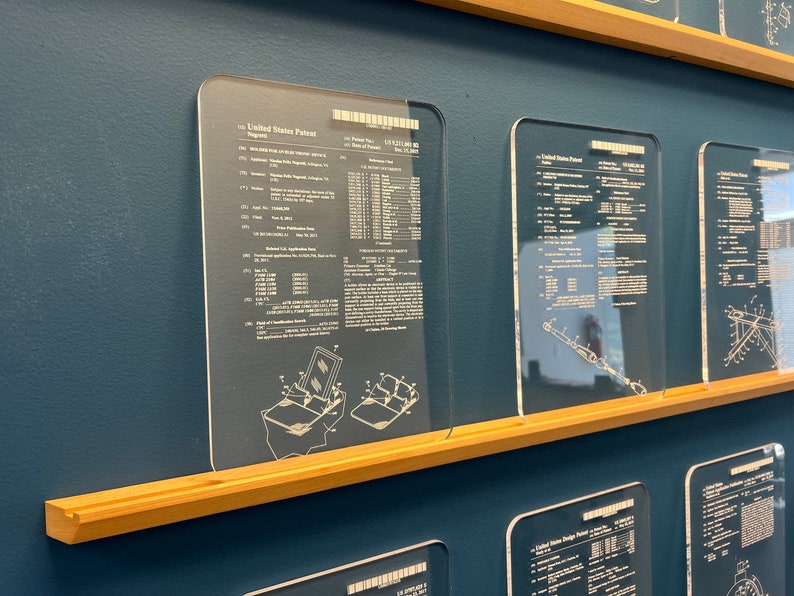 Puede incluir: Una pared con una exposici&oacute;n de patentes de Estados Unidos enmarcadas, impresas en paneles de acr&iacute;lico transparente. Las patentes est&aacute;n montadas en un estante de madera contra una pared azul turquesa.