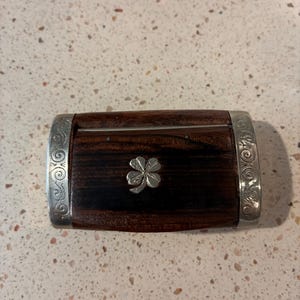 May include: A rectangular wooden box with a dark brown wood grain finish, featuring silver-toned metal end caps with intricate scrollwork engravings and a small silver-toned four-leaf clover emblem centered on the top lid surface.
