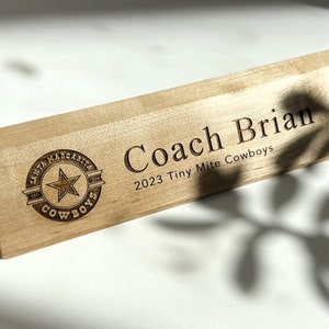 May include: Wooden desk nameplate with the text "Coach Brian" and "2023 Tiny Mite Cowboys" engraved on it. A logo with a star and the text "Santa Margarita Cowboys" is also engraved on the nameplate.