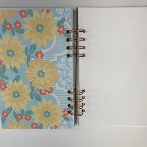 May include: Open notebook with a floral design on the left page and blank white pages on the right. The left page features a light blue background with yellow flowers and red accents. The notebook is bound with gold-colored rings.