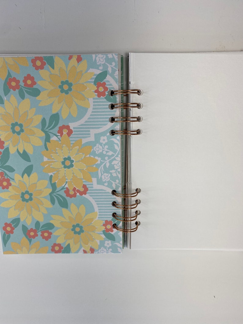 May include: Open notebook with a floral design on the left page and blank white pages on the right. The left page features a light blue background with yellow flowers and red accents. The notebook is bound with gold-colored rings.