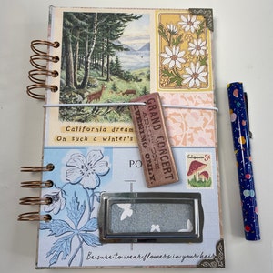 May include: A vintage-style journal with a spiral binding, featuring a forest scene, floral designs, and the text "California dream." Includes a pen with a blue and colorful pattern. The journal has a metal label holder.
