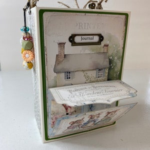 May include: A decorative journal with a vintage aesthetic. The cover features a watercolor illustration of a cottage and the word "Journal" on a metal label. The journal has a beaded charm and a pop-up element with illustrations of mice.