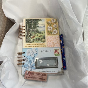 May include: A small, spiral-bound journal with vintage-style illustrations. The cover features a forest scene and floral designs. Additional elements include a decorative label, a pencil, and a tag that reads "GRAND CONCERT". The journal is presented on white tissue paper.