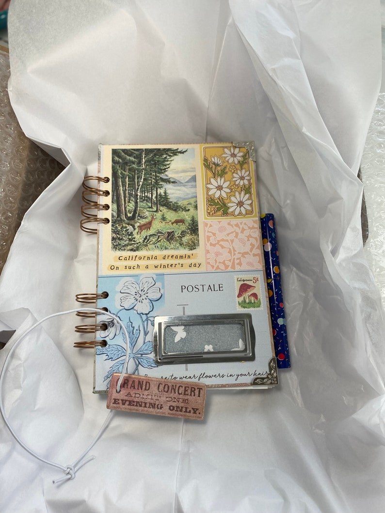 May include: A small, spiral-bound journal with vintage-style illustrations. The cover features a forest scene and floral designs. Additional elements include a decorative label, a pencil, and a tag that reads "GRAND CONCERT". The journal is presented on white tissue paper.