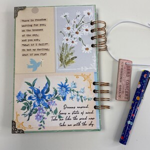 May include: A spiral-bound journal with a floral design and decorative corner accents. The journal features a poem and floral illustrations in blue and white. A blue pen with colorful dots and a small concert ticket are also visible.