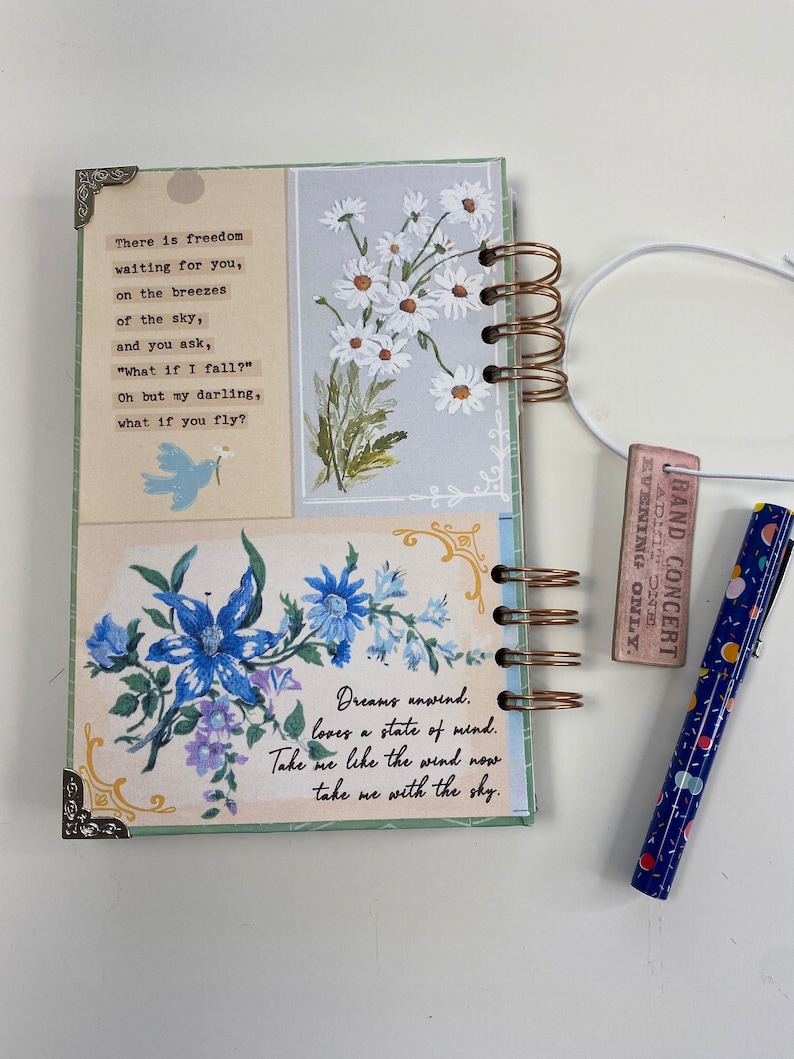 May include: A spiral-bound journal with a floral design and decorative corner accents. The journal features a poem and floral illustrations in blue and white. A blue pen with colorful dots and a small concert ticket are also visible.