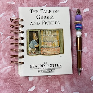 May include: A spiral-bound notebook with the title "The Tale of Ginger and Pickles" by Beatrix Potter. A decorative pen with purple, blue, and white beads sits beside the book on a pink background.