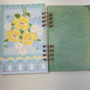 May include: Open journal with a floral design. The left page features a light blue background with yellow flowers, white daisies, and blue accents. The right page has a green chevron pattern. The journal is bound with a gold spiral.