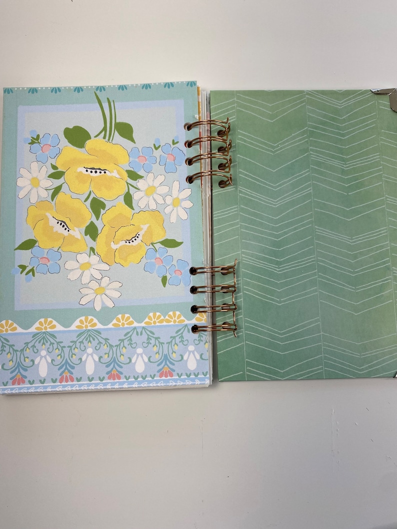 May include: Open journal with a floral design. The left page features a light blue background with yellow flowers, white daisies, and blue accents. The right page has a green chevron pattern. The journal is bound with a gold spiral.