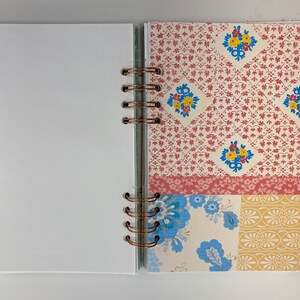 May include: Open journal with white blank pages and a decorative patterned section. The patterned section features a mix of floral and geometric designs in red, blue, and yellow on a cream background. The journal is bound with copper-colored rings.