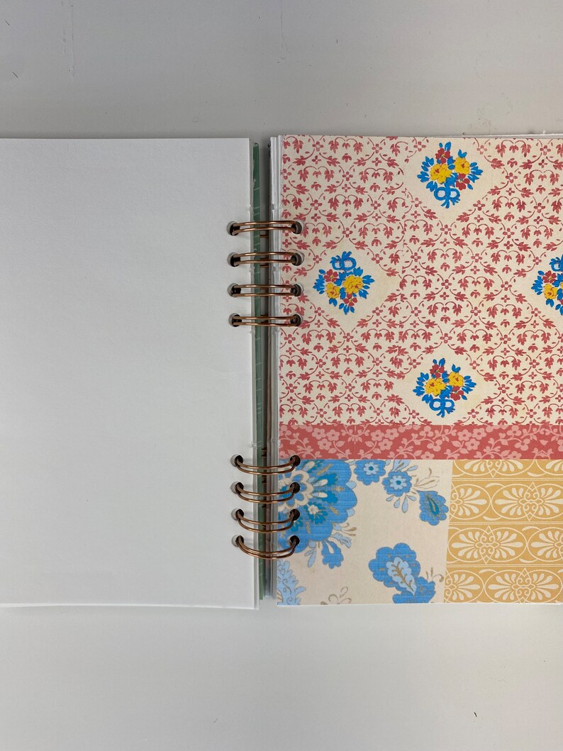 May include: Open journal with white blank pages and a decorative patterned section. The patterned section features a mix of floral and geometric designs in red, blue, and yellow on a cream background. The journal is bound with copper-colored rings.