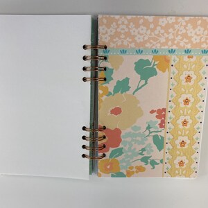 May include: Open notebook with blank white pages and a floral patterned cover. The cover features a mix of peach, yellow, and blue floral designs, with a copper-colored spiral binding. The notebook is open, revealing the blank pages.
