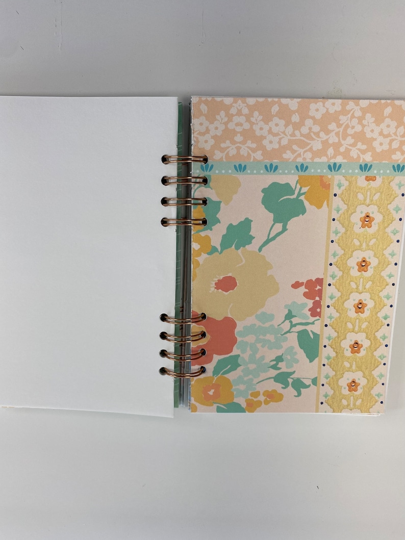 May include: Open notebook with blank white pages and a floral patterned cover. The cover features a mix of peach, yellow, and blue floral designs, with a copper-colored spiral binding. The notebook is open, revealing the blank pages.