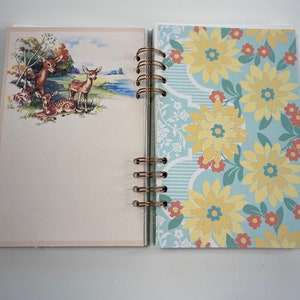 May include: Open journal with a spiral binding. The left page features a vintage illustration of deer near a body of water. The right page has a floral pattern with yellow flowers on a light blue background.