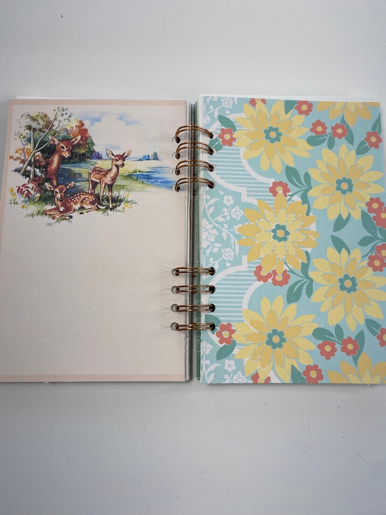 May include: Open journal with a spiral binding. The left page features a vintage illustration of deer near a body of water. The right page has a floral pattern with yellow flowers on a light blue background.
