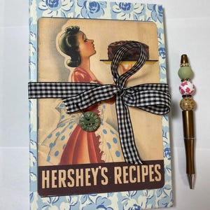 May include: A vintage-style recipe book with a blue floral pattern and a black and white checkered ribbon. The cover features a retro illustration of a woman holding a cake, with "HERSHEY'S RECIPES" printed below. A decorative pen with colorful beads is next to the book.