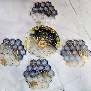 May include: A set of resin coasters with a bee theme. The coasters are in the shape of honeycombs and a round shape. The colors are black, white, and gold with glitter accents. Each coaster features a gold bee design.