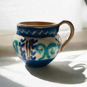 Könnte beinhalten: Handgefertigte Keramiktasse mit rundem Körper und Henkel. Die Tasse hat ein blau-weißes Farbschema mit dekorativen floralen Mustern in Türkis und Dunkelblau. Der Rand hat ein wellenförmiges blaues Design.