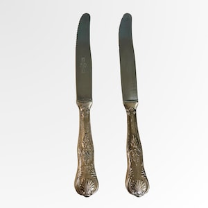 Könnte beinhalten: Zwei silberne Buttermesser mit gezahnten Klingen und verzierten Griffen. Die Griffe weisen ein detailliertes Blumenmuster auf. Die Messer werden vor einem weißen Hintergrund präsentiert, was ihr elegantes, vintage Aussehen hervorhebt.