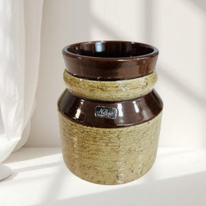 May include: A cylindrical ceramic vessel featuring a two-tone design with a dark brown glazed rim and base, contrasted by a textured, speckled beige middle section. A small rectangular label on the front displays the text Nittsjö Sweden.