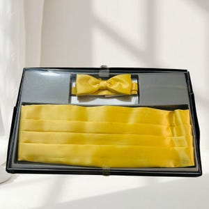 May include: A bright yellow satin cummerbund and a matching pre-tied bow tie presented in a black display box. The cummerbund features horizontal pleats across its surface, while the bow tie is positioned in a separate compartment above it.