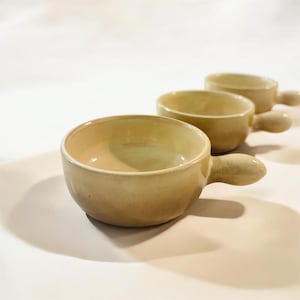 Könnte beinhalten: Drei beige Keramik-Suppenschalen mit Griffen sind diagonal angeordnet. Die Schalen haben eine runde Form und eine glatte, glänzende Oberfläche. Die Griffe sind klein und gebogen. Die Schalen sind leer.