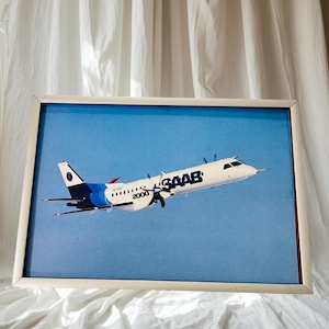 以下が含まれることがあります： 青い空を背景に、白と青のSAAB 2000飛行機が飛行している額入りのプリント。飛行機の側面には「SAAB」の文字が印刷されています。フレームは明るい色です。