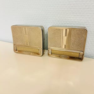 May include: Two vintage silver-toned cigarette cases. Each case has a textured, rectangular design with a central vertical stripe and a hinged lid. The word "RAPID" is embossed on the bottom edge of each case.