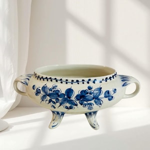 Könnte beinhalten: Eine weiße Keramikschale mit blauen Blumenmustern und zwei Griffen. Die Schale hat drei kleine Füße und einen dekorativen blauen Rand. Das Blumenmuster zeigt stilisierte Blumen und Blätter.