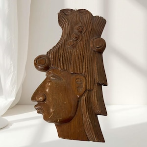 May include: A brown wooden wall plaque carved in the profile of a human head with an elaborate, textured hairstyle featuring circular accents. The carving displays a prominent nose, defined lips, and a stylized ear, with a smooth, polished finish.