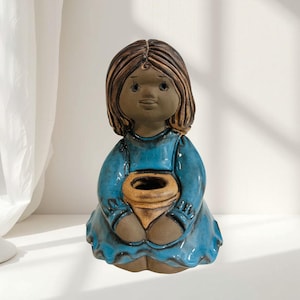 May include: A ceramic figurine of a seated girl with brown hair and a blue dress. The figurine is holding a tan-coloured pot. The girl's face has a serene expression. The figurine is a decorative item.