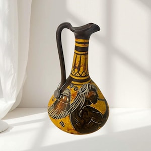 May include: A ceramic pitcher featuring a dark brown handle and neck with a yellow body. The vessel is decorated with black hand-painted figures and geometric patterns. A small circular tag hangs from the handle by a thin silver-colored string.