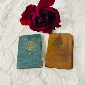 Puede incluir: Dos pequeños libros antiguos, uno verde azulado y otro marrón, se exhiben con una rosa roja oscura sobre una superficie de encaje blanco. El libro verde azulado tiene texto dorado y un diseño de sol con el año 1922. El libro marrón tiene el año 1923.