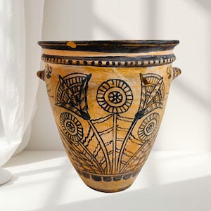May include: A tan ceramic vase featuring black floral patterns. The vase has a wide opening, two small handles, and a black rim. The floral design incorporates circular and fan-shaped motifs, giving it a vintage appearance.