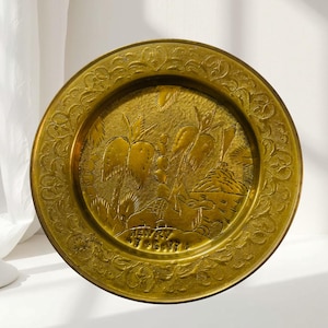 May include: A round brass decorative plate featuring an embossed tropical scene with palm trees and a camel. The center includes the engraved text "HENRY" above the date "7-6-57". The wide rim is decorated with a detailed, repeating floral pattern.
