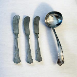 Può includere: Un set di posate d'epoca, tra cui tre coltelli da burro e un mestolo. I coltelli da burro hanno lame curve e manici decorati. Il mestolo ha una coppa rotonda e un lungo manico curvo. Tutti i pezzi sono color argento.