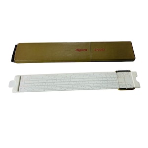 Könnte beinhalten: Ein weißer Rechenschieber mit schwarzen Markierungen und einer braunen Pappschachtel. Auf der Schachtel stehen die Worte "ARISTO STUDIO" in Rot. Es ist ein Vintage-Recheninstrument.