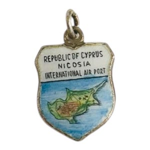 Op de afbeelding: Een zilverkleurige bedel in de vorm van een schild, met de tekst "REPUBLIC OF CYPRUS NICOSIA INTERNATIONAL AIR PORT." De bedel heeft een blauwe achtergrond met een kaart van Cyprus in groen en bruin.